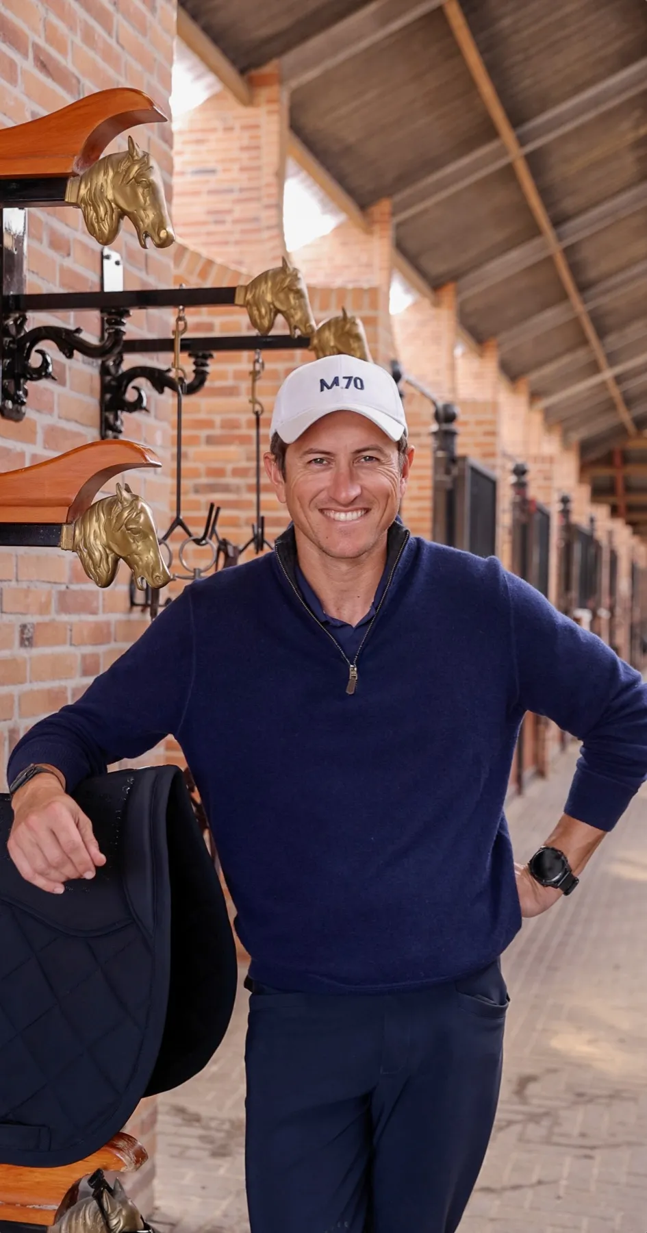 Sean Crooks in the barn aisle, leaning on a saddle and smiling in an M.70 cap